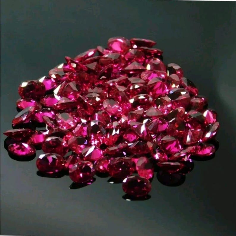 May include: A pile of faceted, deep red gemstones. The gems are scattered across a reflective black surface, catching the light and displaying multiple facets. The stones vary in size and shape, creating a sparkling, luxurious display.