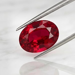 以下が含まれることがあります： シルバーカラーの金属製ピンセットで保持された、鮮やかなオーバルカットの赤いルビーの宝石。宝石の表面はファセットカットされており、光を反射して、透明で磨かれた構造全体に深く飽和した深紅の色合いを映し出しています。
