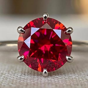 May include: A ring featuring a large, round-cut, vibrant red gemstone set in a polished silver-toned metal band. The stone is secured by six prongs, showcasing a faceted surface that reflects light with deep crimson hues and sharp geometric patterns.