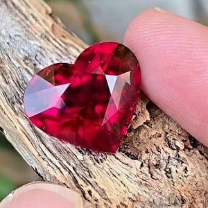 May include: A faceted, heart-shaped gemstone in a deep red hue. The gem is held against a piece of weathered wood, showcasing its brilliant cut and reflective surfaces. The gemstone's facets catch the light, creating a sparkling effect.