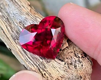 Puede incluir: Una gema facetada en forma de corazón, de un tono rojo intenso. La gema se sostiene contra un trozo de madera envejecida, mostrando su talla brillante y superficies reflectantes. Las facetas de la gema capturan la luz, creando un efecto brillante.