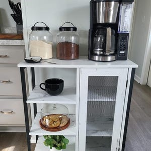 May include: A white and black kitchen cabinet with a coffee maker, glass jars, and a black coffee cup. The cabinet has shelves with a glass dome with cookies and a small plant. The text "TrendyvogueArt" is visible.