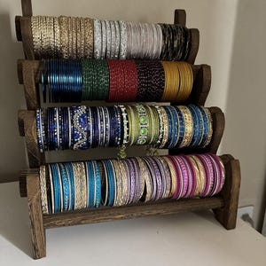 May include: A wooden display stand holding multiple rows of colorful bangles. The bangles are in various colors, including gold, silver, black, blue, green, red, and pink. The text "TrendyvogueArt" is visible.