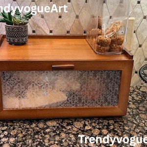 May include: A wooden bread box with a clear, patterned glass front and a wooden top. A small potted plant sits on the top, and a clear container with baked goods is on the right. The text "TrendyvogueArt" is visible.