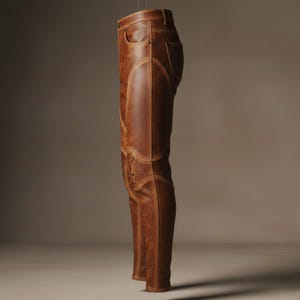 K&ouml;nnte beinhalten: Braune Lederhose mit dekorativen N&auml;hten und Schn&uuml;rdetails am Unterschenkel. Die Hose hat ein klassisches F&uuml;nf-Taschen-Design und ist vor einem neutralen Hintergrund aufgeh&auml;ngt.