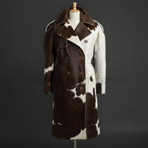 May include: A long, double-breasted coat with a striking cowhide pattern. The coat features a split design, with one side in brown and white cowhide and the other in solid white. It has a notched collar and multiple buttons.