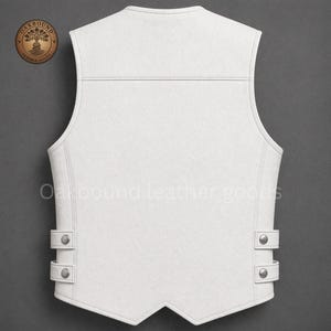 May include: A white leather vest with a V-neck design. The vest features two sets of adjustable straps with silver-colored buttons on the sides. The text "Oakbound leather goods" is printed on the vest.