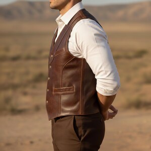 May include: A brown leather vest with a classic design. The vest has a button-up front, a pocket, and a tailored fit. The vest is worn over a white dress shirt and brown pants. The background is a desert landscape.