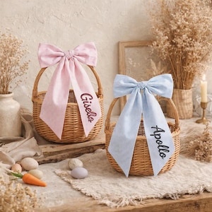 Puede incluir: Dos cestas de mimbre tejidas con grandes lazos a rayas. Un lazo es rosa con el nombre "Gisella" bordado en cursiva. El otro lazo es azul claro con el nombre "Apollo" bordado en cursiva. Las cestas están sobre una superficie de madera.