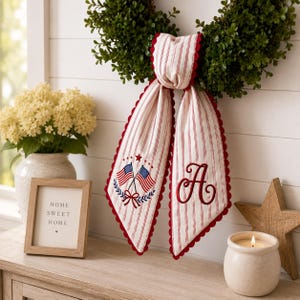 Puede incluir: Una cinta decorativa de tela con rayas verticales rojas y blancas y un borde festoneado rojo. Un lado muestra un dise&ntilde;o bordado de dos banderas estadounidenses cruzadas con un lazo, mientras que el otro lado presenta una letra A bordada.