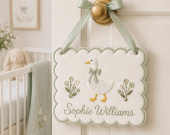 May include: A white, scalloped-edge sign with a duck and floral embroidery, hanging from a brass doorknob with a sage green ribbon. The sign reads "Sophie Williams" in script. A framed picture and crib are in the background.