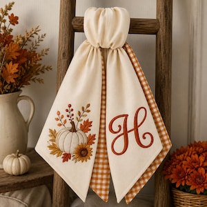 May include: A hanging kitchen towel featuring a cream-coloured fabric with an embroidered white pumpkin, autumn leaves, and a sunflower on one side, and a large embroidered orange letter H on the other, with orange and white gingham trim.