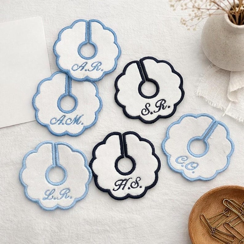 May include: White and blue embroidered coasters with scalloped edges. Each coaster features a central hole and a vertical slit, with initials embroidered in blue thread. The coasters are arranged on a light-colored surface.