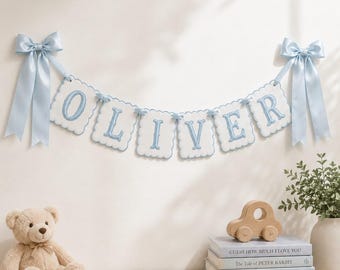 May include: A baby name banner spelling "OLIVER" in blue embroidered letters on white scalloped flags, suspended by blue bows. A teddy bear, wooden car, and books are on a wooden surface below.
