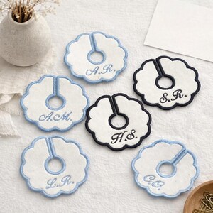 May include: Embroidered white and blue coasters with a unique design. Each coaster features a scalloped edge and a central opening, with initials embroidered in a delicate script. The coasters are arranged on a light-colored surface.