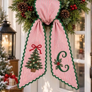 Peut inclure: Une suspension de porte en tissu décoratif avec des rayures verticales rouges et blanches et une bordure festonnée vert foncé. Un côté présente un sapin de Noël brodé avec un nœud rouge, l'autre côté affiche une grande lettre C verte.
