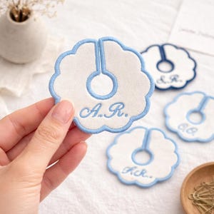 May include: Embroidered white and light blue floral-shaped garment clips. The clips have a light blue border and a central opening. The initials "A.R." are embroidered in light blue on one clip. Other clips with different initials are also visible.
