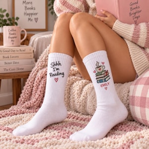 May include: A pair of white crew socks featuring graphic designs. The left sock displays the black text Shhh I'm Reading with a small red heart. The right sock features a stack of books topped with a coffee mug and the name EMILY in red.