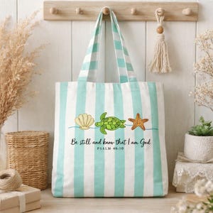 May include: A white tote bag with vertical stripes in light blue. The bag features a seashell, turtle, and starfish design, with the text "Be still and know that I am God" and Psalm 46:10.