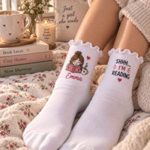 May include: A pair of white ribbed crew socks with ruffled cuffs. The left sock features a cartoon illustration of a person reading a book with a mug and the name Emma. The right sock displays the text SHHH... I'M READING with small red hearts.