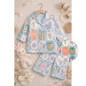 May include: Light blue Easter-themed pajama set with an egg pattern featuring bunnies, flowers, and bows. The set includes a button-down top and matching pants. A plush bunny and decorative elements complete the scene.
