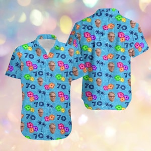 Può includere: Camicia azzurra con bottoni con un motivo ripetuto del volto di un uomo, il numero 70, fiori tropicali e palme. La camicia ha maniche corte e un colletto classico.