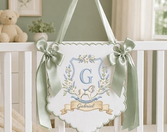 May include: A white fabric wall hanging with scalloped edges, featuring an embroidered blue shield, a letter G, a rocking horse, and floral accents. The name Gabriel is stitched on a tan banner. It has sage green satin ribbons for hanging.