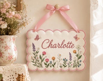 May include: A rectangular wall hanging with scalloped edges, featuring the name Charlotte embroidered in pink script. The cream-colored fabric is decorated with colorful floral embroidery and two small butterflies, suspended by a pink satin ribbon.