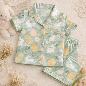 Puede incluir: Un conjunto de pijama verde menta con un estampado de conejos blancos y huevos de Pascua. La parte superior de manga corta y los pantalones cortos tienen ribetes amarillos. El conjunto est&aacute; hecho de una tela suave y ligera, ideal para primavera o Pascua.