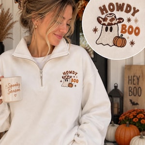 May include: A white quarter-zip sweatshirt featuring an embroidered design on the left chest. The embroidery depicts a ghost wearing a brown cow-print cowboy hat next to a pumpkin, with the text "HOWDY BOO" and small star accents in brown.