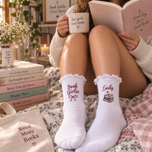May include: A pair of white crew socks with ruffled cuffs. The left sock features the text Book Girlie in maroon with a bow graphic. The right sock features the name Emily in maroon above a stack of three books with a small heart graphic.