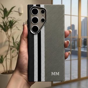 Puede incluir: Una funda de teléfono con un diseño de rayas grises, negras y blancas. La funda tiene una superficie texturizada y presenta un recorte para la cámara con múltiples lentes. Las iniciales "MM" están impresas en la esquina inferior derecha.