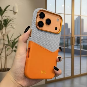 Op de afbeelding: Een tweekleurige telefoonhoes in oranje en grijs, met een gouden camerakader. Het bovenste gedeelte is grijs geweven stof, de onderkant is oranje. De initialen "KK" zijn goudkleurig in de rechter benedenhoek geëmbosseerd.