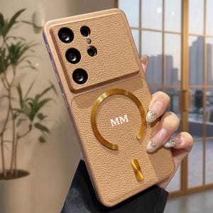 Puede incluir: Una funda de teléfono color beige con textura similar al cuero, que presenta un anillo magnético circular dorado y un pequeño detalle vertical en oro. Incluye un módulo de cámara elevado y las letras MM impresas en el centro del anillo.