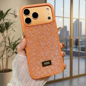 May include: Orange and glitter iPhone case with a rectangular design. The case has a glittery texture on the back and a solid orange frame. The camera area is also orange. A small, rectangular metal plate with the letters "MM" is on the lower back.