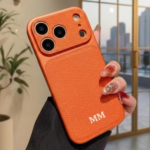 Puede incluir: Funda de teléfono naranja texturizada con un módulo de cámara y las iniciales "MM" en blanco. La funda tiene un aspecto similar al cuero y está diseñada para proteger las lentes de la cámara del teléfono. La funda se sostiene en una mano con uñas cuidadas.