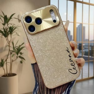 May include: A gold glitter phone case with a gold camera lens cover. The case has the name "Cecana" written in cursive. The phone case is held in a person's hand.