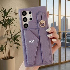 May include: A lilac phone case with a cardholder and a hand strap. The case has a textured finish and gold-coloured hardware. The initials "MM" are printed on the cardholder.