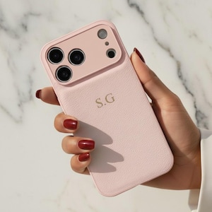 Può includere: Custodia per telefono rosa chiaro con finitura strutturata, con modulo fotocamera e le iniziali "S.G.". La custodia è tenuta in mano con smalto rosso, su sfondo marmoreo.