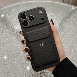 Peut inclure: Coque de téléphone noire avec une finition texturée et une poche pour cartes. L'étui comporte un module caméra avec trois objectifs et un flash. Les initiales "A.S" sont imprimées en or sur le porte-cartes.
