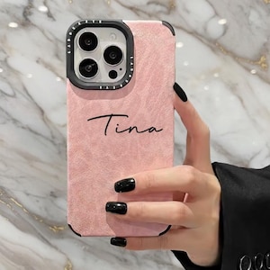 Può includere: Una custodia per telefono rosa con il nome "Tina" in corsivo. La custodia ha una superficie strutturata e un bordo nero attorno alla fotocamera e ai bordi. La custodia del telefono è tenuta in una mano con smalto nero.