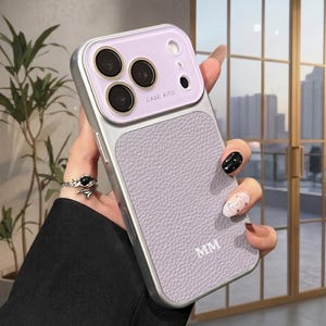 May include: A lavender and silver phone case with a textured leather-like back and a protective camera lens cover. The case has the text "CASE AITO" and initials "MM".