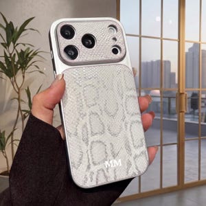 Peut inclure: Une coque de smartphone avec un motif texturé en peau de serpent blanc et gris clair. La coque présente un cadre en argent métallique et une zone de découpe pour l'appareil photo avec un motif assorti. Les lettres MM sont imprimées en blanc.