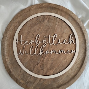 Könnte beinhalten: Ein kreisförmiges Schild aus hellem Holz mit dem lasergeschnittenen Schriftzug "Herbstlich Willkommen" in Schreibschrift. Das Schild ist von einem dünnen Holzring umrahmt und liegt auf einem größeren, runden Holztablett mit sichtbarer Maserung.