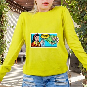 May include: A bright yellow long-sleeve sweater with a comic-style patch. The patch features a woman spraying a bottle labeled "Anti-Cupid Spray" at a cartoon Cupid, with the words "Begone, Pest!"