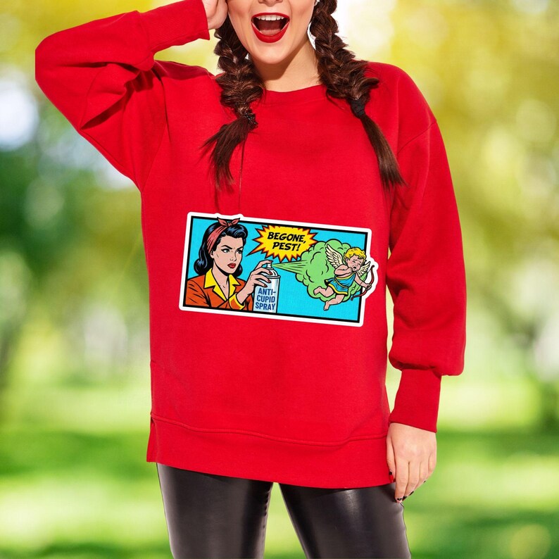 May include: A red sweatshirt with a comic book-style graphic. The graphic features a woman spraying "Anti-Cupid Spray" at a cartoon Cupid with the words "Begone, Pest!" in a speech bubble. The sweatshirt has long sleeves.