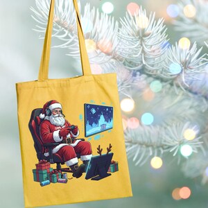 Puede incluir: Bolso tote amarillo mostaza con un Papá Noel de dibujos animados jugando. Papá Noel lleva auriculares y sostiene un mando, con una escena invernal pixelada en la pantalla. El bolso tiene un tema festivo con cuernos de reno y regalos envueltos.