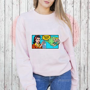 May include: A light pink sweatshirt featuring a pop art style graphic. The graphic depicts a woman spraying "Anti-Cupid Spray" at a cartoon cupid with the text "Begone, Pest!" in a yellow burst.