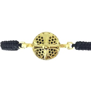May include: Gold-coloured bracelet with a circular, cross-shaped centrepiece and a honeycomb design. The bracelet has black braided cords on either side, connected by gold clasps. The jewellery is set against a white background.