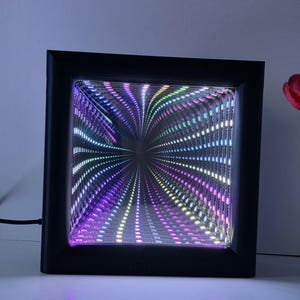 May include: A black square LED infinity mirror with a mesmerizing display of colorful lights. The lights radiate from the center, creating a tunnel effect with shades of purple, green, and white. The frame is a simple, modern design.